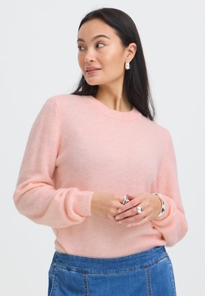 Femme portant un pull rose doux et un jean bleu, avec des boucles d'oreilles, des bagues et un bracelet en argent, regardant sur le côté, les mains jointes.