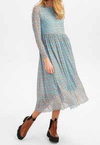 Modré květinové šaty s dlouhými rukávy, s překládaným pasem a volnou sukní. Mají kulatý výstřih a průhlednou látku. Spárovány s hnědými kotníkovými botami.