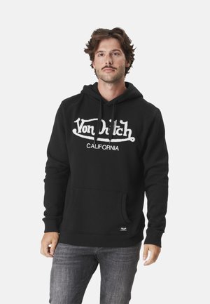 Homme aux cheveux ondulés portant un sweat à capuche noir avec le texte « Von Dutch California », associé à un jean gris, se tenant devant un fond uni.