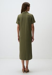 Abito midi verde oliva, a maniche corte, con taglio dritto e cucitura posteriore. Tessuto morbido; indossato con sandali beige.