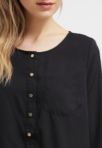 Blusa nera con scollo rotondo, chiusura frontale con bottoni in oro e una sola tasca sul petto. Tessuto morbido e leggero.