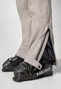Lower legs wearing beige ski pants with side zippers partially open over black HEAD ski boots secured with buckles.