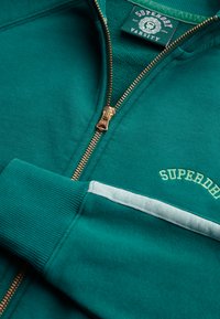 Felpa con zip color verde petrolio, con polsini a coste, piccolo ricamo con logo e una striscia contrastante di colore azzurro lungo la zona delle tasche. Dettaglio cerniera dorata.