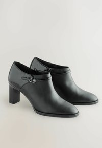 Bocanci negri din piele la gleznă cu o bordură din piele lucioasă, vârf ascuțit, toc pătrat de 6 cm și curea decorativă cu detaliu cu cataramă argintie.
