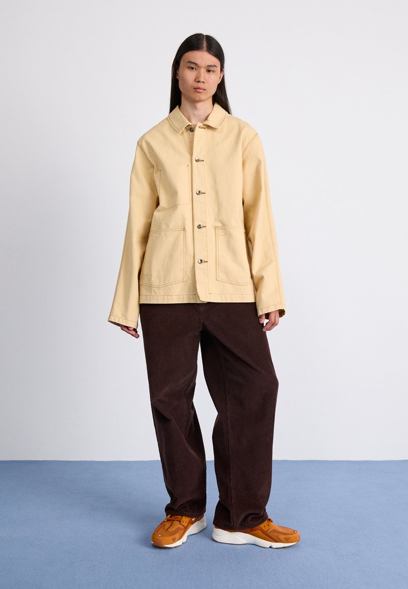 Chaqueta de color amarillo claro con cuello y botones plateados, combinada con pantalones de pana marrón oscuro y zapatillas naranjas sobre un suelo azul.