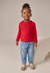 Rød langærmet skjorte med flæser, blå denim jeans og brune sko med leopardmønster. Stående positur mod en neutral baggrund.