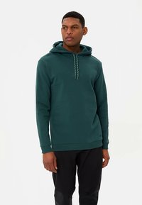 Grüner Baumwoll-Hoodie mit Kordelzugkapuze, Kängurutasche und gerippten Bündchen, getragen mit schwarzer Hose. Glatte Textur, lässige Passform.