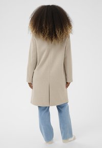 Cappotto lungo beige realizzato in tessuto soffice, con uno spacco sul retro, maniche inserite e una silhouette dritta, abbinato a jeans a gamba larga di un azzurro chiaro.