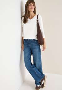 Top bianco a maniche lunghe con scollatura a V, jeans blu a gamba larga e sneakers marroni. La vestibilità è comoda, il tessuto è morbido e le sneakers presentano dettagli bianchi.