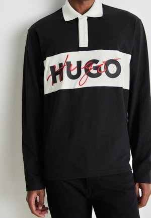 Black long-sleeve polo shirt with white collar and chest stripe featuring black and red "HUGO" text worn by person.