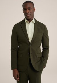 Saco de traje ligero de color verde oscuro con un corte ajustado, dos bolsillos frontales y cierre de un solo botón, combinado con una camisa de color verde claro con cuello.