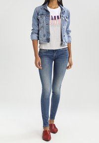 Chaqueta de denim, de color azul claro, ajustada con dos bolsillos frontales. Camiseta gris con diseño de bloque de colores. Jeans skinny azules. Zapatillas rojas tipo slip-on.