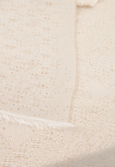 Υφασμα σε κρεμ απόχρωση με λεπτές χρυσές κλωστές και κρόσσια κατά μήκος μιας ακμής, το οποίο παρουσιάζεται σε στρωμένα πτυχώματα.