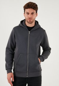 Siva zip-up hoodie s prednjim džepom, podesivom kapuljačom i rebrastim manžetama. Mekani materijal od pamuka i opušteni kroj.