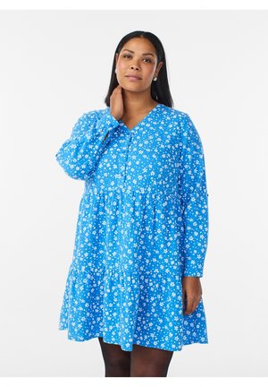 Ung kvinde med langt mørkt hår iført en blå blomstret langærmet kjole, står med den ene hånd på sin nakke mod en hvid baggrund.