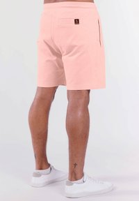 Helle rosa Shorts aus weichem Material, ausgestattet mit einer Gesäßtasche und einem kleinen Logostick. Kombiniert mit weißen Sneakers mit grauen Akzenten.