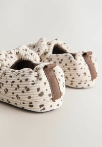 Des chaussons en tissu crème doux avec des taches léopard marron et des tirettes marron à l'arrière, posés sur une surface claire.