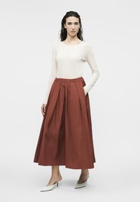 Femme debout portant un haut blanc ajusté à manches longues, une jupe midi plissée couleur rouille, et des mules blanches à bout pointu avec des talons kitten.