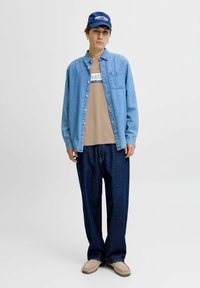 Denimskjorte i lyseblå, beige grafisk t-shirt, løse mørkeblå jeans og en marineblå kasket med hvid tekst, med afslappet pasform og bløde teksturer.