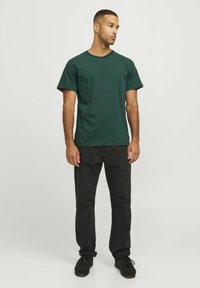 T-shirt di cotone verde scuro con collo a giro, maniche corte e vestibilità rilassata, abbinata a jeans neri e scarpe nere.
