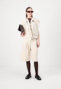 Beige mouwloze jas met ritsen en zakken, bruine top, crèmekleurige shorts, zwarte panty, en zwarte instappers, met een donkere clutch in de hand.