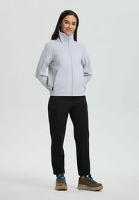 Woman standing with hands behind back wearing light gray zip-up jacket, black pants, and brown hiking shoes with blue laces on plain background.