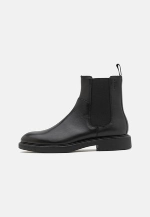 Botte de cheville en cuir noir avec une texture lisse, des panneaux latéraux élastiques et une languette. Bout rond et semelle en caoutchouc plate avec un minimal de relief.