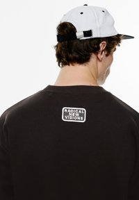 Sort sweatshirt med ribbede manchetter og krave, udstyret med et hvidt broderet logo "RADICAL NEW VISIONS" på ryggen. Akkompagneret af en hvid kasket.