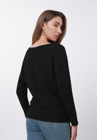 Juodas, siūluotas ilgomis rankovėmis viršus su plačiu kaklu, derinamas su mėlynais džinsais. Audinys atrodo tekstūruotas ir prigludęs prie juosmens.