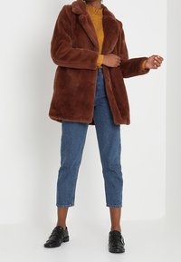 Abrigo de piel sintética marrón con cuello en pico, llevado sobre un top de punto amarillo. Combinado con jeans de cintura alta azules y zapatos negros.