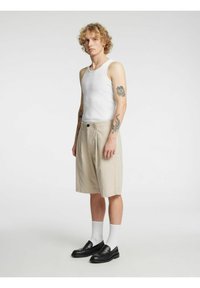 Beige stripete shorts med folder, kombinert med en hvit tanktopp og svarte loafers. Modellen har krøllete hår og tatoveringer.