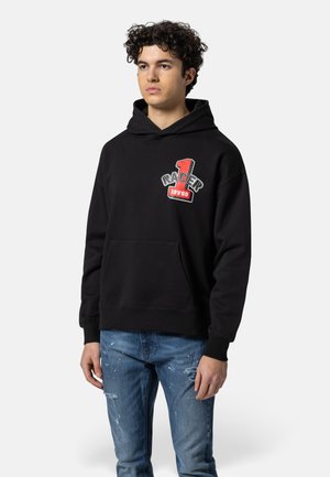 Junger Mann mit lockigem Haar, der einen schwarzen Hoodie mit rotem und weißem "Racer 1969"-Logo und ausgewaschene blaue Jeans trägt, vor einem schlichten Hintergrund steht.