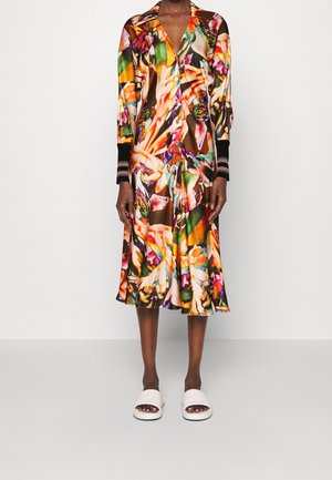 Frau trägt ein farbenfrohes, knöpfbares Midikleid mit Blumenmuster, schwarzen gestreiften Manschetten und weißen offenen Slides, steht vor einem einfarbigen Hintergrund.