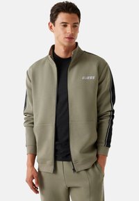 Chaqueta verde oliva con cremallera, paneles laterales negros, logotipo en el pecho, puños acanalados y bolsillos, hecha de tela suave, combinada con una camisa negra.