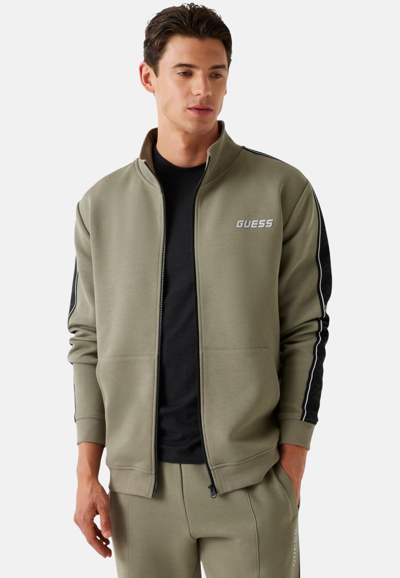 Chaqueta verde oliva con cremallera, paneles laterales negros, logotipo en el pecho, puños acanalados y bolsillos, hecha de tela suave, combinada con una camisa negra.