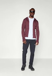 Felpa bordeaux con zip sopra una t-shirt bianca, pantaloni sportivi neri e scarpe nere, indossata da una persona con occhiali da sole, in piedi davanti a uno sfondo neutro.