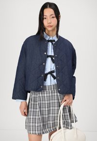 Marineblauwe gewatteerde denimjack met drukknoopjes, lichtblauw rimpelshirt en grijs geruit geplooid rokje. Model houdt een witte handtas vast.
