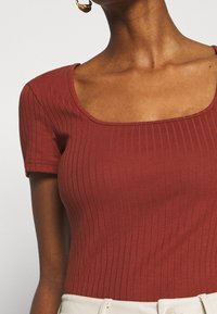 Top acanalado de color óxido con escote cuadrado y mangas cortas, confeccionado en tejido texturizado, combinado con una cintura de color claro.