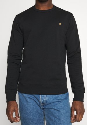Homme portant un sweatshirt noir uni à manches longues avec un petit logo "F" jaune sur la poitrine et un jean bleu, sur fond blanc.