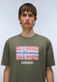 Camiseta de algodón verde oliva con un gráfico de la bandera noruega y líneas onduladas en rojo y azul, con "NAPAPIJRI geographic" impreso debajo.