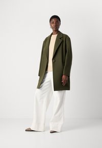 Olive green wool blend coat with a notched lapel, single button closure, and side pockets, paired with a cream sweater and wide white trousers.