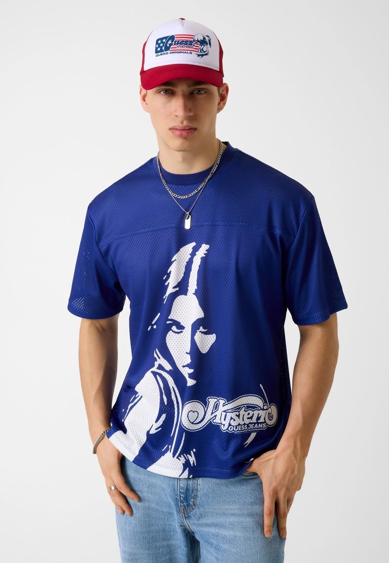 Νέος άνδρας που φορά μπλε γραφικό δίκτυο T-shirt με ένα άσπρο στυλιζαρισμένο πρόσωπο και κείμενο, ανοιχτό μπλε τζιν, κόκκινο και άσπρο καπέλο και ασημένια αλυσίδα κολιέ.