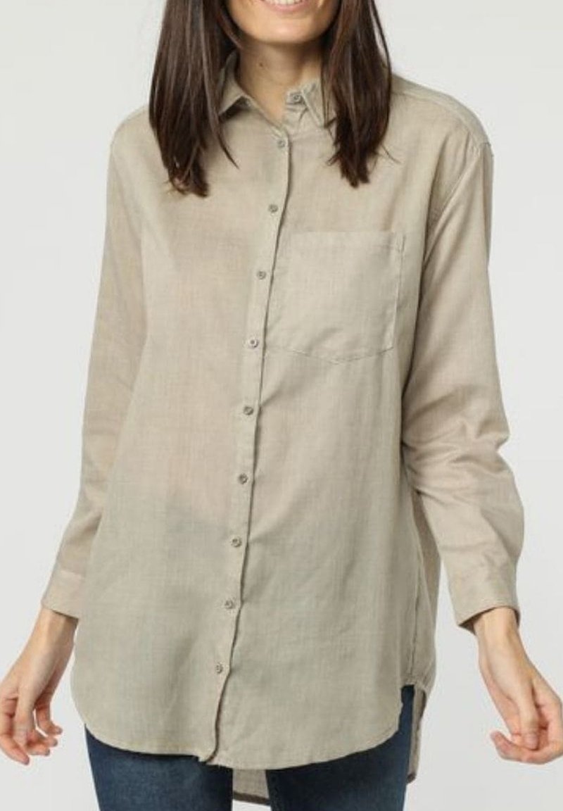 Mujer que lleva una camisa beige suelta abotonada con mangas largas y un bolsillo en el pecho, combinada con vaqueros azul oscuro sobre un fondo liso.