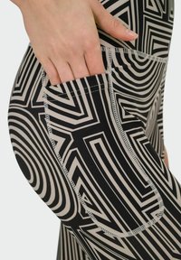 Leggings en negro y beige con un patrón geométrico, con franjas prominentes y un bolsillo lateral visible. Detalles de tejido suave y elástico.