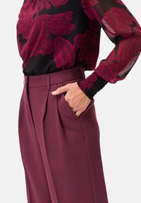 Pantaloni a gamba larga bordeaux con pieghe, abbinati a una blusa nera con motivi floreali magenta e maniche trasparenti. Polsini neri texture.