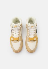 Tenisky Nike Air Trainer mají béžový a krémový svrchní materiál s žlutými akcenty, texturované překryvy, bílé tkaničky a suchý zip pro dodatečnou podporu.