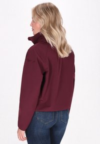 Burgundinės minkštosios striukės su aukštu apykakle, pasižyminčios liekna forma ir lygiu paviršiumi, derinamos su mėlynomis džinsinėmis kelnėmis.