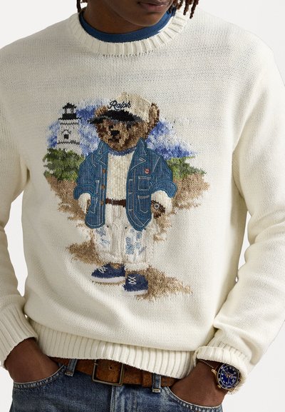 Horgolt krém színű pulóver, kék dzsekis és sapkás medve hímzéssel. Bordás mandzsetták és alsó szegély, farmernadrággal és órával viselve.