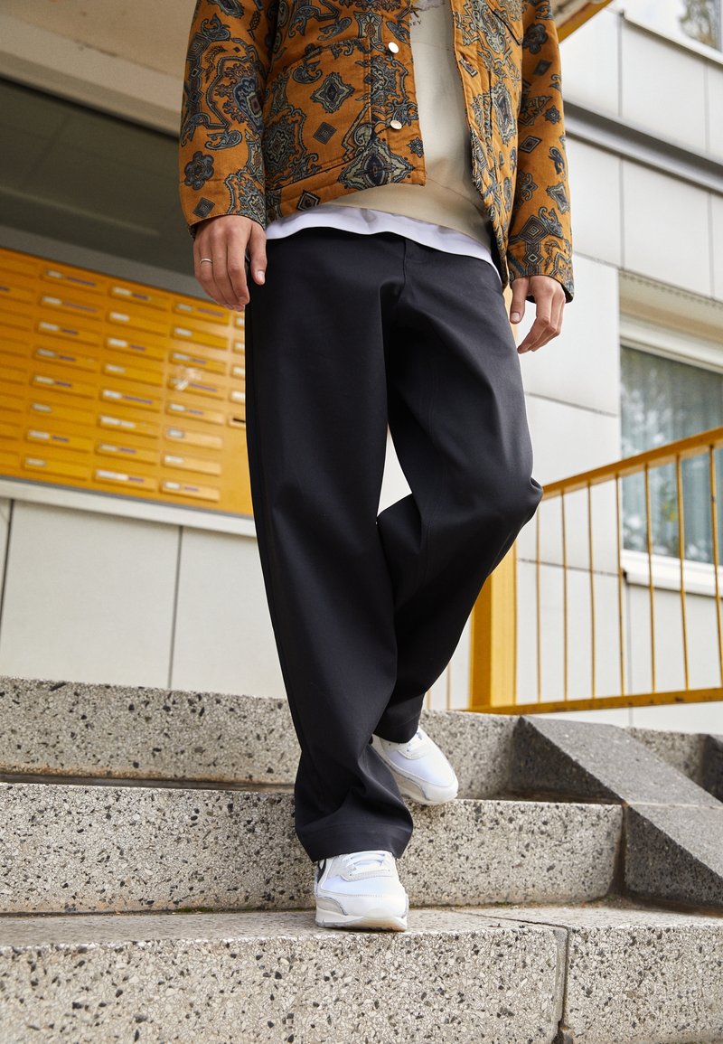 Person som bär en mönstrad orange jacka, svarta vida byxor och vita sneakers, går ner för betongtrappor utanför en byggnad.