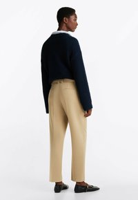 Marineblauwe gebreide trui, beige op maat gemaakte broek en zwarte instapschoenen, met daaronder een overhemd met kraag. Zachte texturen en gestructureerde pasvorm.